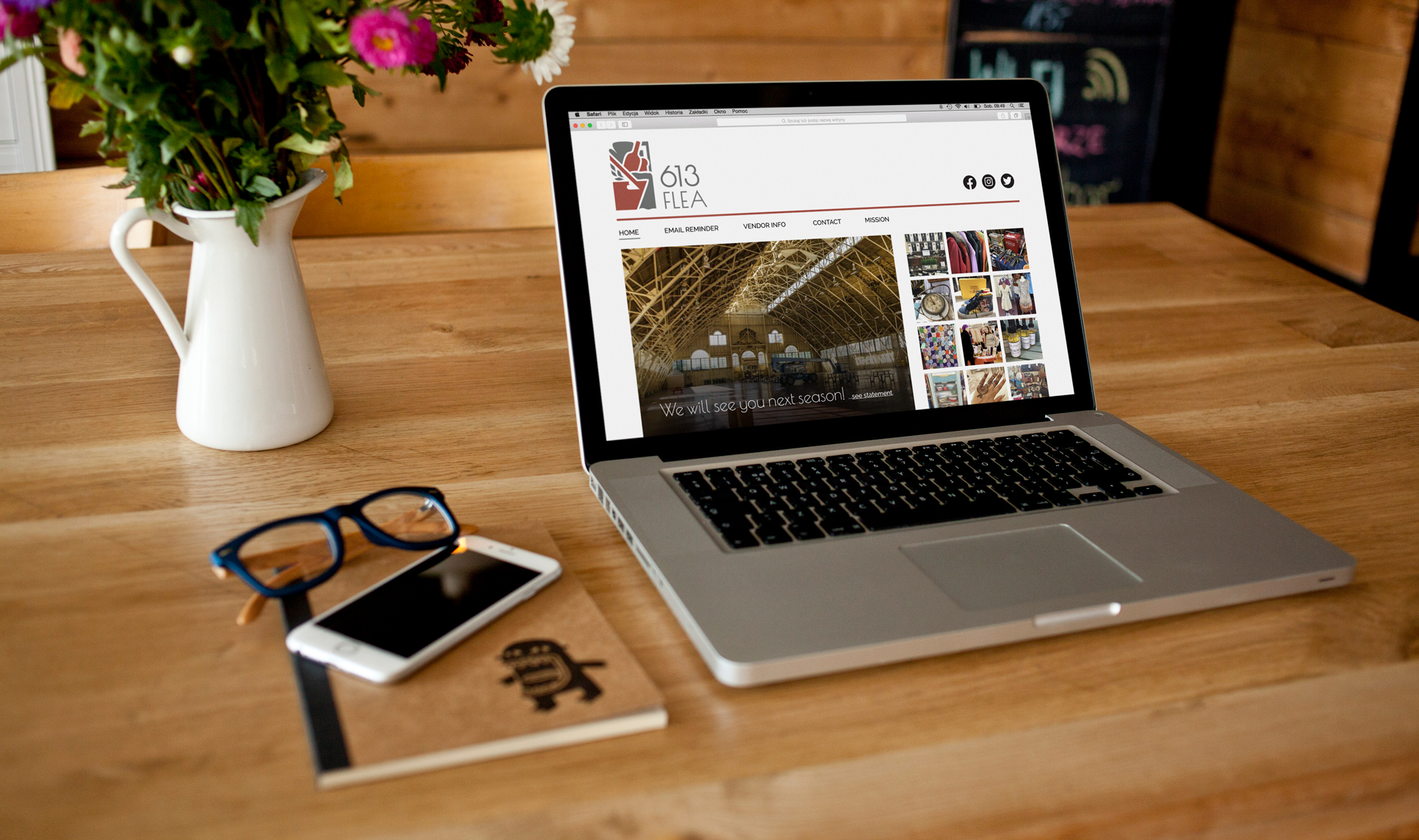 A laptop displays a mockup of the 613 Flea website homepage, on a wood surface with a white of pink flowers, a notepad, pair of glasses and smartphone.