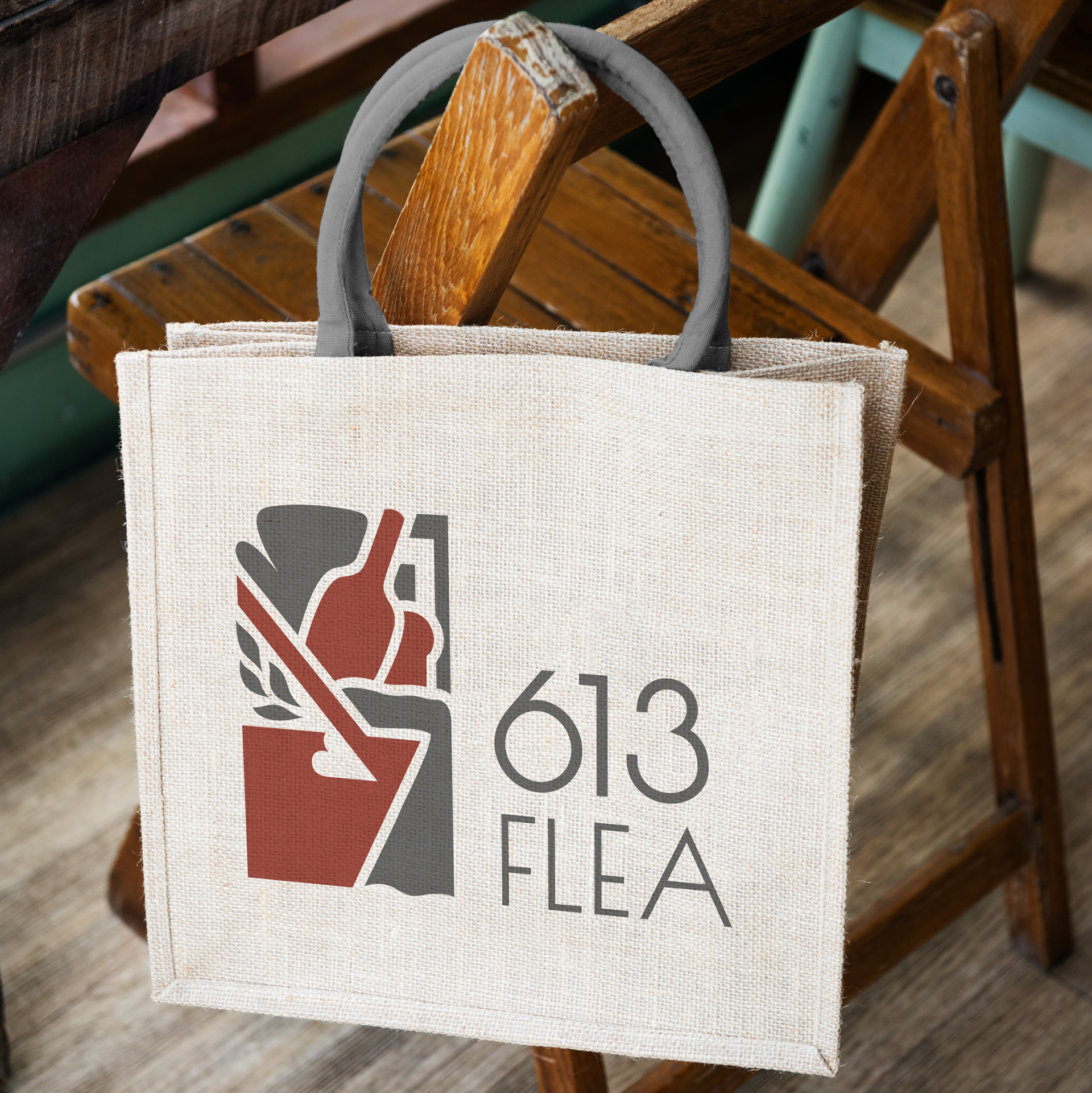 A wicker looking totebag with the logo on it, with a grey handle, hangs on a wooden slat chair.